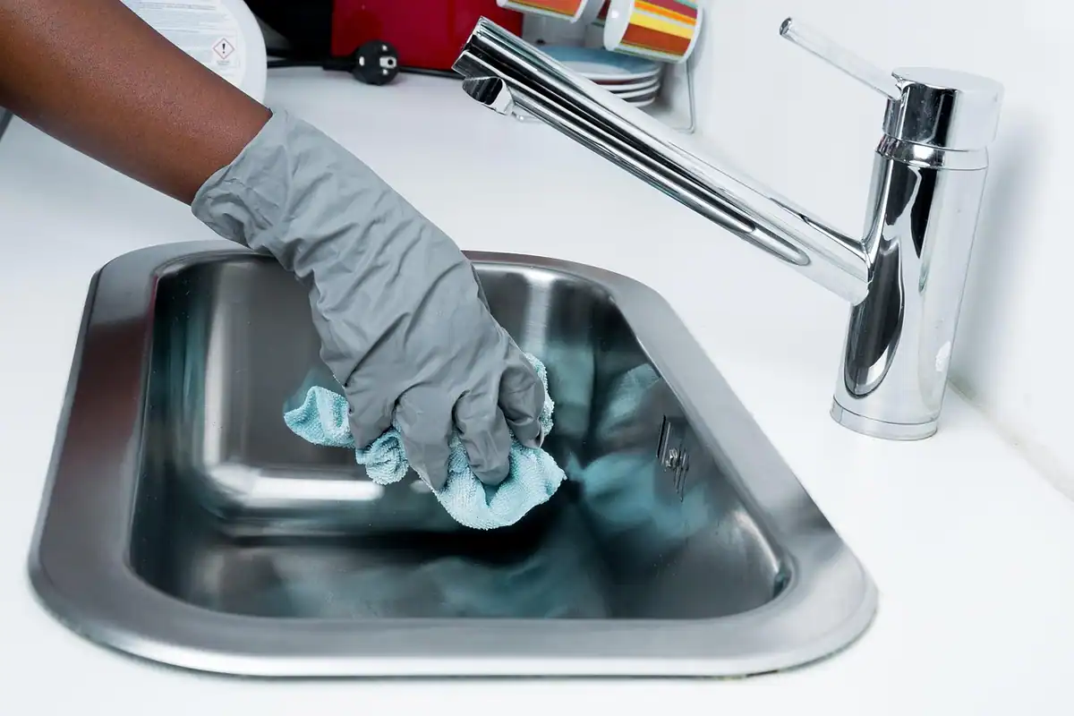 Mână cu mănușă gri care șterge chiuveta de inox din bucătărie